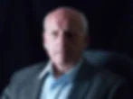 Serious bald older man in a gray blazer and white shirt, seated against a dark studio background, lit from the left.