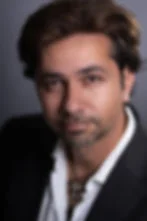 Close-up portrait of a man in a black jacket and white shirt, looking seriously at the camera against a gray background.