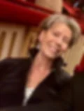 Smiling woman with short gray hair in a black jacket sitting in a theater. Red seats and beige panels in the background.