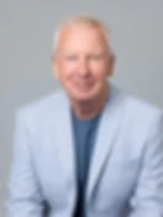 A man smiles, wearing a light blue blazer and blue shirt against a gray background, conveying a friendly, professional mood.